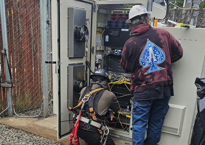 Two electricians working in an electrical panel below a cell tower.