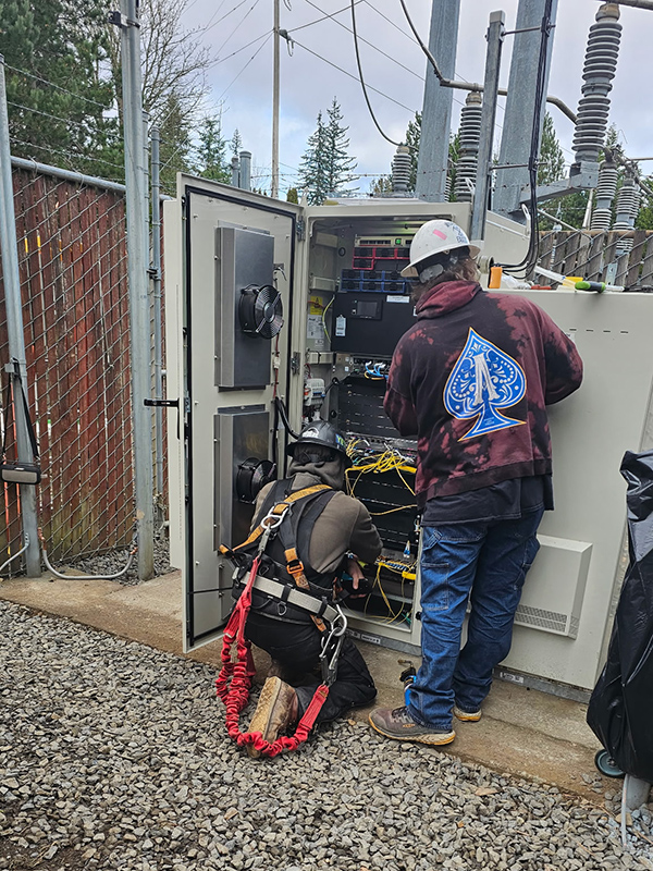 Two electricians working in an electrical panel below a cell tower.