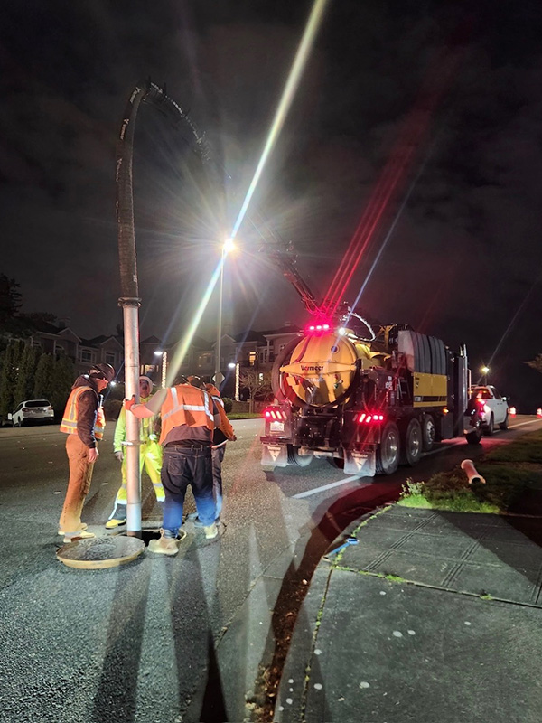 Large truck with a vacuum hose inserted in a manhole with workers around it.